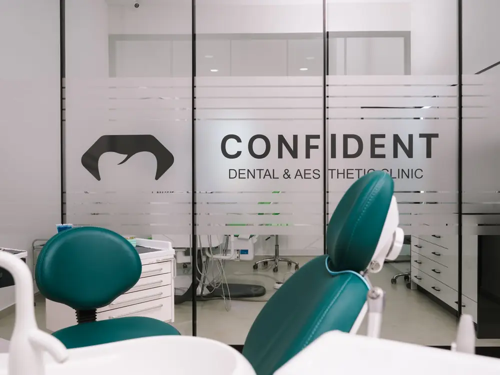 Pogled sa tirkizne stomatološke stolice prema staklenom zidu s logom Confident Dental & Aesthetic Clinic